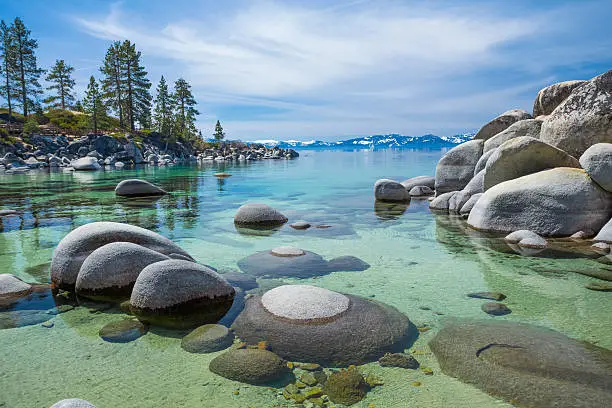 Picture of shallow calm waters of Lake Tahoe near San Harbor State Park. There are some clouds in the sky and rocks seeing through shallow transparent water. There are pine trees on the left side near horizon line and snowy mountain peaks of Sierra Nevada.