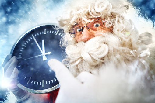 Closeup of funny Santa Claus holding big round clock and pointing to it because it's 5 to 12 and is almost Christmas time. It's snowing.He's looking at camera with surprised expression and raised eyebrows. Low angle view, wide shot.