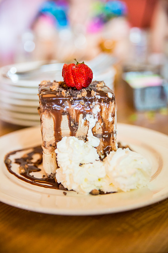 A yummy piece of coffee ice cream cake sits on a plate. There is chocolate sauce on the cake. There is whipped cream on plate and a strawberry on top. It's being served for dessert to people who are blurred in the background. Taken with Canon 5D Mark 3. rm