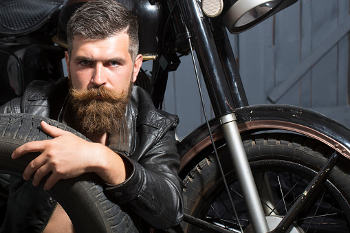 Handsome unshaven male biker in leather jacket sitting near motorcycle in garage with big black rubber spare wheel looking forward on wooden wall background, horizontal picture
