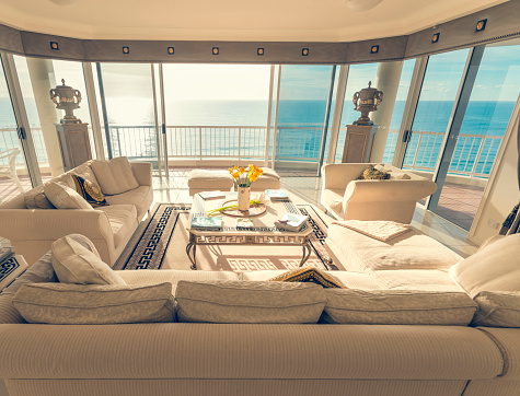 Living room in a luxury beachfront apartment with view. There is a magnificent view of the ocean from the floor to ceiling windows. The living room has a large sofa, coffee table and some flowers. Very spacious and luxurious.