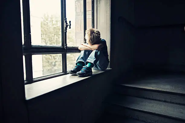 Depressed 6 years old child sitting on window in old staircase. The boy is crying hiding his head in arms. 