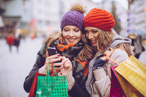 Female friends shopping in city. Holding shopping bags and using smart phone. Wearing warm clothes, hat and scarf. Vienna, Austria. Day time.