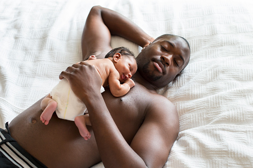 A barechested male lying down with his newborn sleeping baby on his chest.