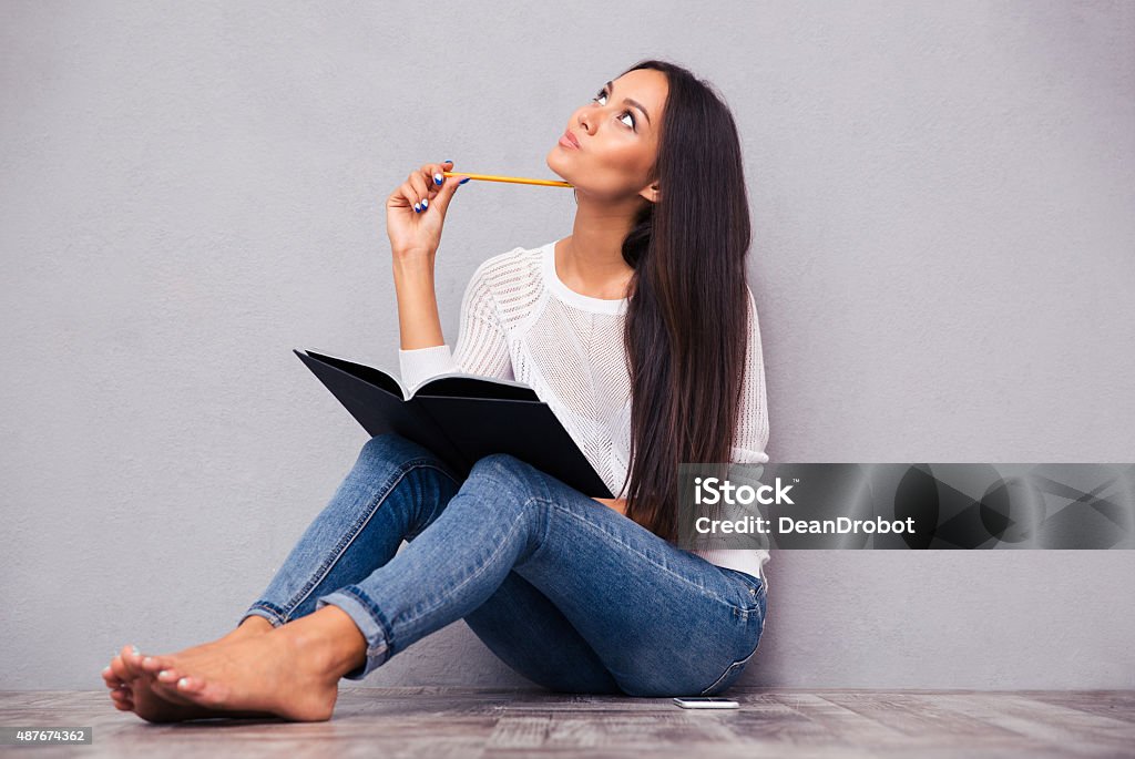 Girl sitting on the floor with notepad and pencil Portrait of a pensive girl sitting on the floor with notepad and pencil on gray background 2015 Stock Photo