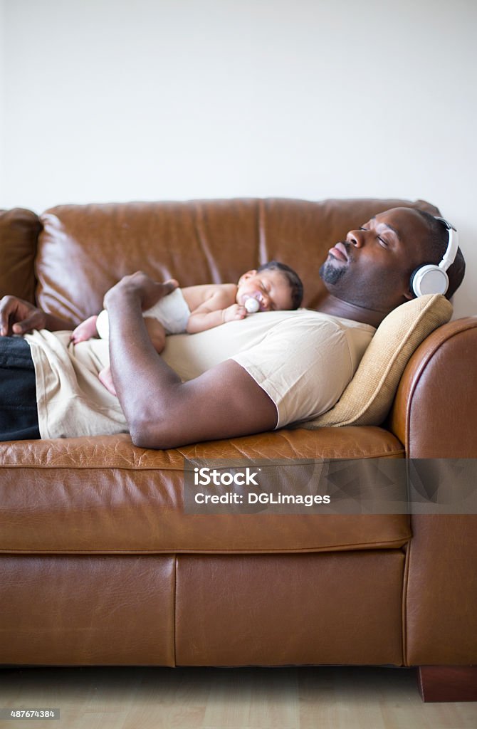 Daddy and Daughter at home Father at home with his newborn baby daughter sleeping on the sofa. He has headphones in and is wearing casual clothing. Sleeping Stock Photo