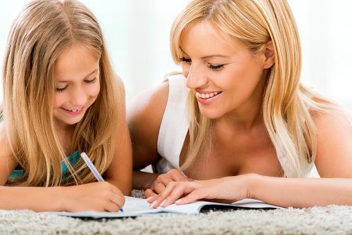 Smiling mid adult mother helping her daughter with her homework. They are lying o the carpet and writing.