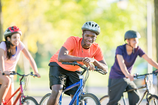 A multi-ethnic group of high school age friends are going on a bike ride at the park together on a beautiful sunny day. One man is smiling and looking at the camera.