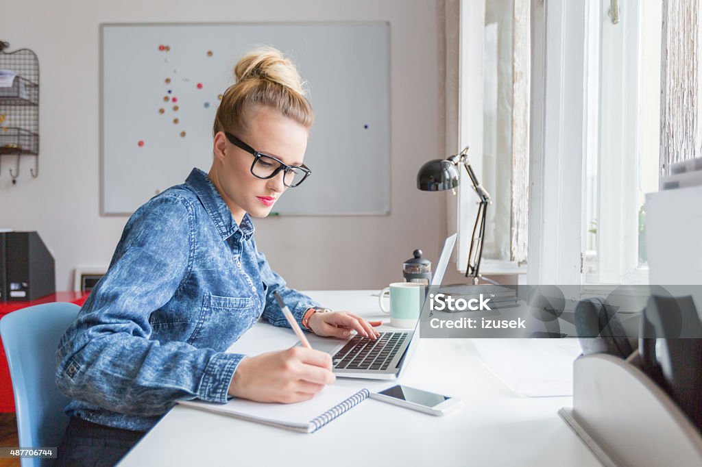 Woman working on computer in an office Blonde woman wearing jeans shirt and nerd glasses sitting at the desk in an office and working on laptop.  Marketing Stock Photo