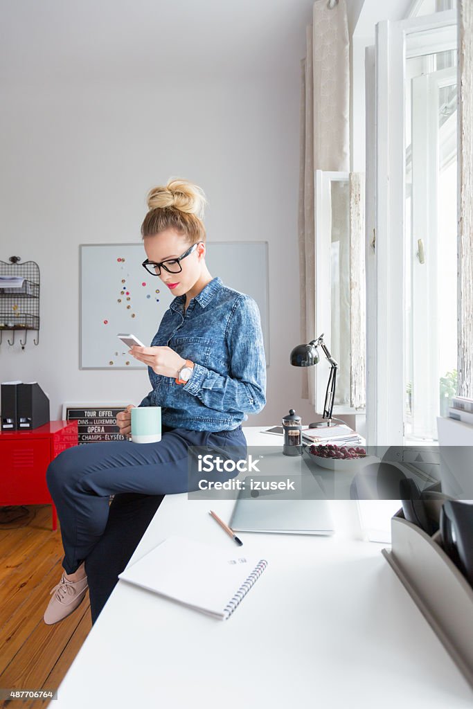 Woman texting on smart phone in an office Blonde woman wearing jeans shirt and nerd glasses sitting on the desk in an office and texting on smart phone, holding cup of coffeee in hand. 2015 Stock Photo