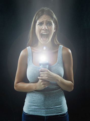 A young brunette holding a flashlight while looking scared and isolated on a black background