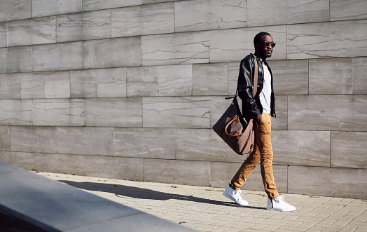 Fashion stylish young african man in sunglasses and black rock leather jacket with bag walking over grey textured city background