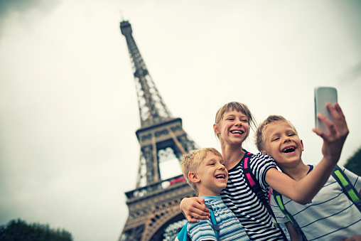 Little girl with her younger brothers are taking selfie near The Eiffel Tower. The tower is visible in the background. Cloudy day. The kids aged 9 and 6 are hugging and smiling at the mobile phone.Paris, France.