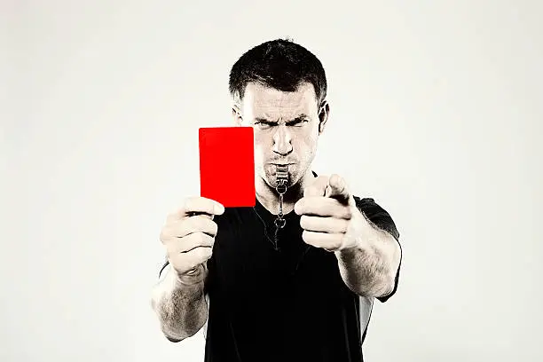 A male, Caucasian sports referee showing a red penalty card and blowing a whistle. Shot in the studio against a light background and with a retro photoshop effect.