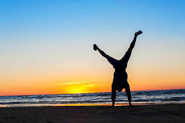 Young sporty man making handstand on the beach. Multicolored sunset background. Horizontal summertime outdoors image.
