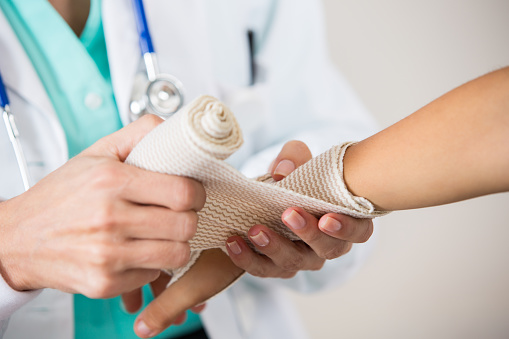 Pediatrician is using an elastic bandage to wrap the sprained wrist of child patient. Doctor is unrolling bandage around little girl's arm. Physician is wearing a lab coat and stethescope. Doctor and patiet's hands are focus of image.