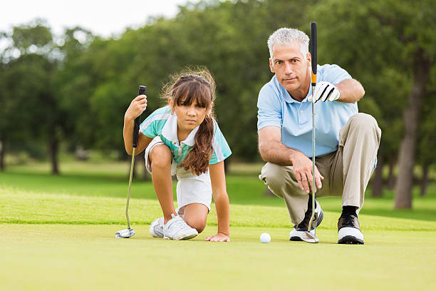 Golf instructor teaching technique to little girl Senior adult Caucasian man is golf pro at country club. He is giving private golf lesson to elementary age Hispanic little girl. He is teaching student how to line up golf ball with putter on green course. golf glove stock pictures, royalty-free photos & images