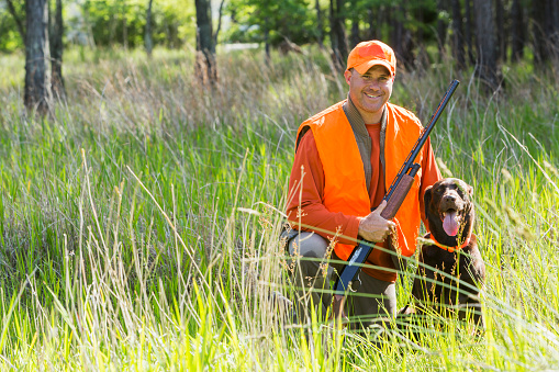 Hunter kneeling on the ground in tall grass, smiling at the camera holding a shotgun.  His hunting dog, a brown labrador retriever, is sitting next to him.  He is wearing an orange safety vest.