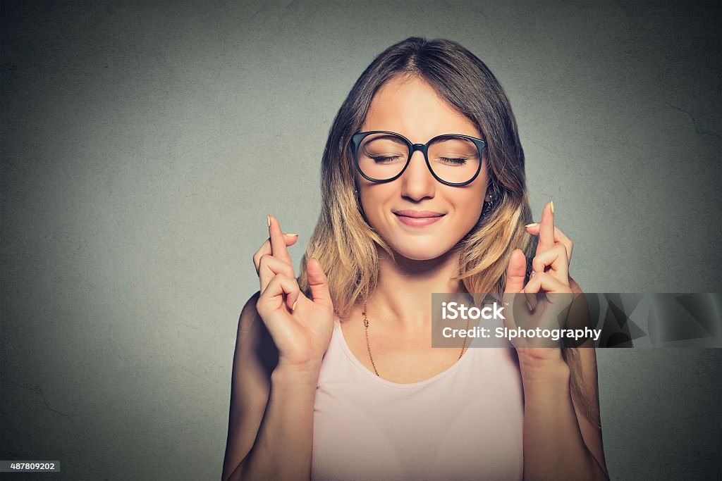 hopeful woman crossing her fingers, eyes closed Closeup portrait hopeful beautiful woman crossing her fingers, eyes closed, hoping, asking best isolated on gray wall background. Human face expression, emotions, feeling attitude reaction Educational Exam Stock Photo