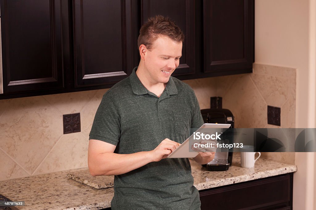 Casual man on his digital tablet in home kitchen. Casual, mid-adult man using his digital tablet at home in kitchen. 30-39 Years Stock Photo