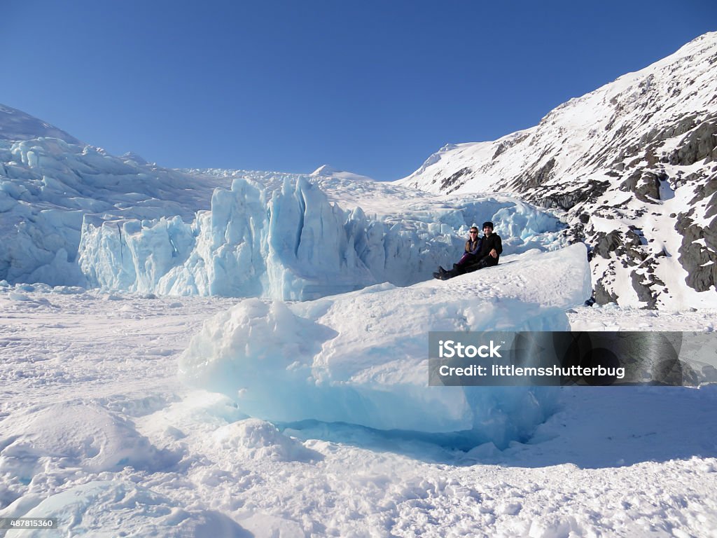 Sitting on an Iceberg from Portage Glacier Two people sitting on top of an iceberg frozen in Portage lake with the Portage Glacier in the background. The shear size of the glacier & iceberg make the people look so tiny. Alaska - US State Stock Photo