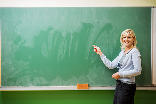 Happy female teacher pointing on a school blackboard and looking at the camera. Copy space.