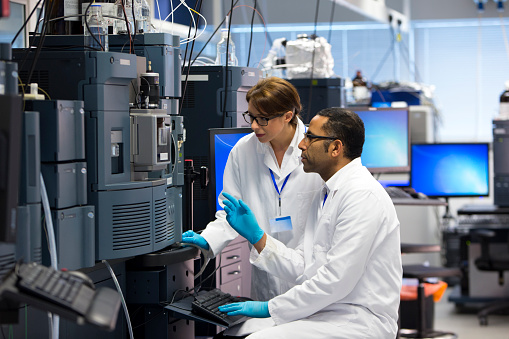 Scientists in a laboratory recording data. Both working beside a special piece of machinery called a Mass spectrometry (MS) , an analytical chemistry technique that helps identify the amount and type of chemicals present in a sample by measuring the mass-to-charge ratio and abundance of gas-phase ions