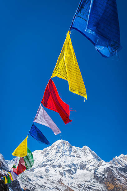 Traditional colourful Buddhist prayer flags flying from the shrine at Annapurna Base Camp in the clear blue high altitude skies over the snow capped summit of Annapurna South (7219m) deep in the Himalayan mountain wilderness of the Annapurna Conservation Area, Nepal. Shallow depth of field with focus on the red flag in foreground. ProPhoto RGB profile for maximum color fidelity and gamut.