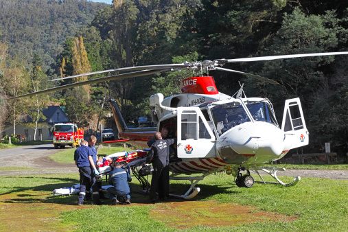 Walhalla, Australia - April 25, 2014: The crew of the Air Ambulance Victoria helicopter Helimed 1 supervise the transfer of an accident victim from ambulance to helicopter on the Anzac Day holiday in a small remote hillside town in rugged countryside.  The ambulance paramedics and doctor assist loading the stretcher onto the waiting chopper.  In the background, the local volunteer fire brigade officer has stationed the fire truck to protect the helicopter landing pad