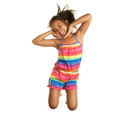 An adorable seven year old Filipino girl on a white background.  She is jumping and is caught in mid air.  She is smiling and having fun.  She is wearing a brightly colored romper