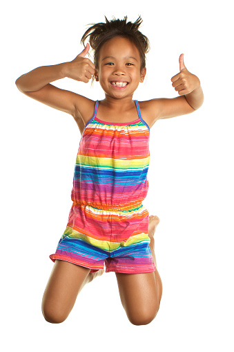 An adorable seven year old Filipino girl on a white background.  She is jumping and is caught in mid air.  She is smiling and giving thumbs up.  She is wearing a brightly colored romper
