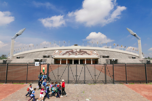 Mexico City, Mexico - July 24, 2013: A group of kids is leaving the Mexico City Olympic Stadium facade. Made for the 1968 Olympics, at the grounds of the Mexico National Autonomous University UNAM, it has a stone mural by artist Diego Rivera. Today is the house of the Pumas professional soccer team.
