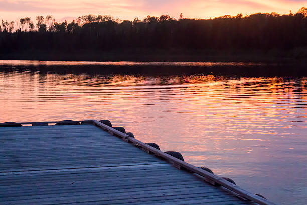 Sunset image on a Canadian lake from a wooden dock.  The water appears pink, reflecting the colorful sky.  The trees on the opposite side of that lake are black and silhouetted.
