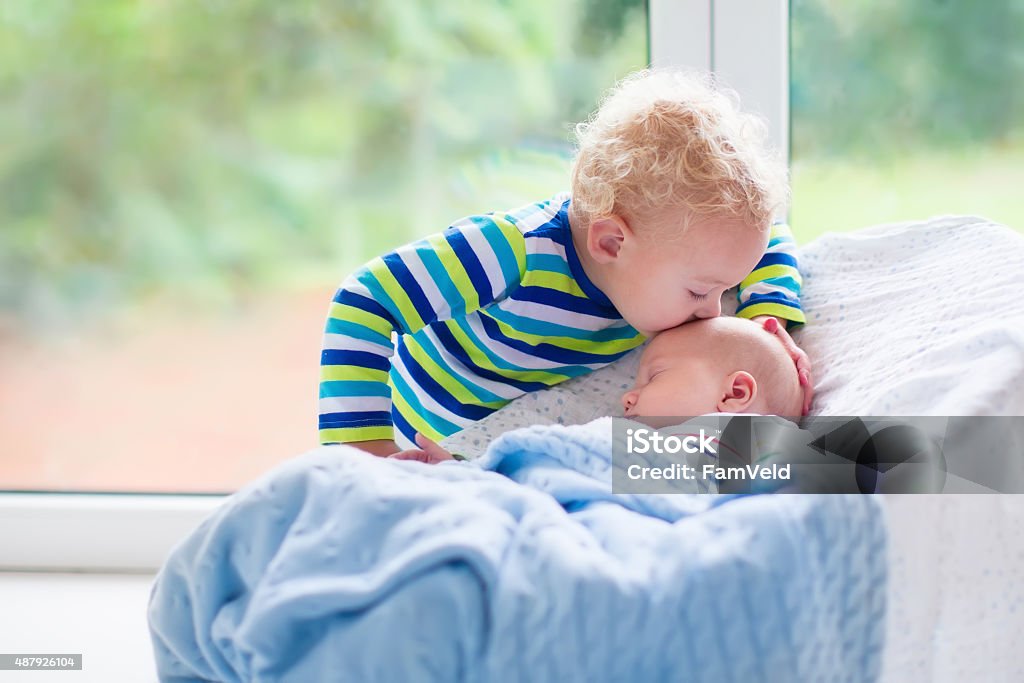 Little boy kissing newborn baby brother Cute little boy kissing his newborn brother. Toddler kid meeting new born sibling. Infant sleeping in white bouncer under a blanket. Kids playing and bonding. Children with small age difference. Baby - Human Age Stock Photo
