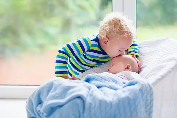 Cute little boy kissing his newborn brother. Toddler kid meeting new born sibling. Infant sleeping in white bouncer under a blanket. Kids playing and bonding. Children with small age difference.