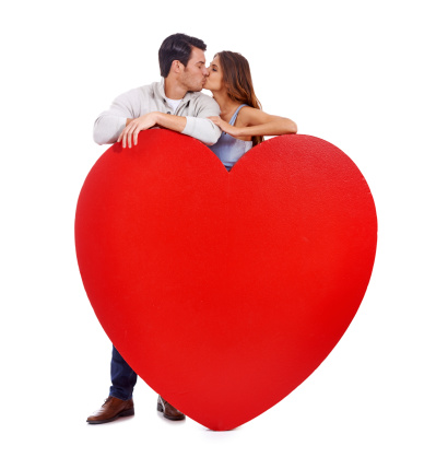 Studio shot of a couple kissing while standing behind a large heart isolated on white