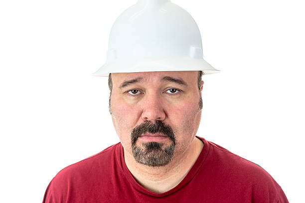Morose glum looking man in a hardhat Morose glum looking man with a goatee beard wearing a hardhat looking at the camera with lacklustre eyes and a depressed expression, isolated on white lazy construction laborer stock pictures, royalty-free photos & images