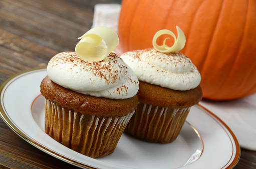 Freshly baked pumpkin cupcakes with cream cheese frosting topped with white chocolate curls, with a seasonal pumpkin in the background.