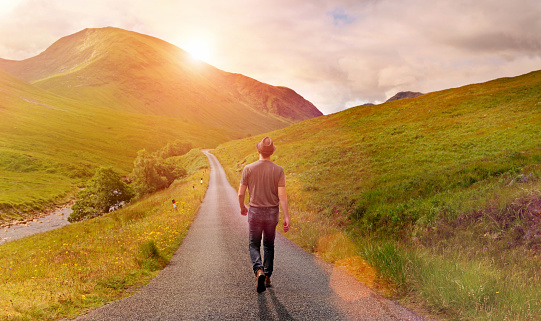 Man with casual clothes walking alone on a narrow country road through a green valley sorrounded by mountains, heading toward the rising sun. Warm sunlight with lens flare. Taking decisions and choosing a direction for new beginnings, following the way forward, not looking back.