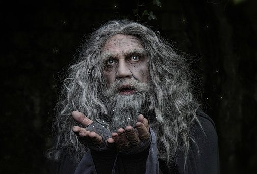 mature man dressed as wizard casting a light spell. Model has long grey unkempt hair and beard, his hands are open as if spreading lights around, his eyes are looking upwards.