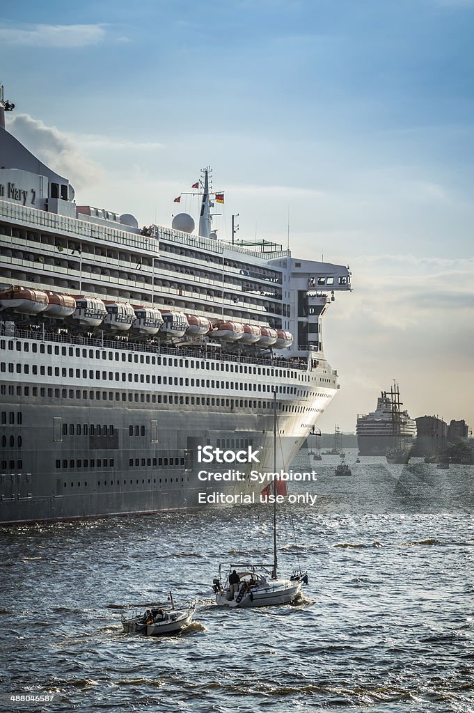 Cruise Ship Queen Mary 2 Hamburg, Germany - May 12, 2013: Criuse Ship Queen Mary 2 is leaving the harbor of Hamburg. Starting on the river Elbe heading to the North Sea. Some small ship are following. People are watching and waving.  Blue Stock Photo