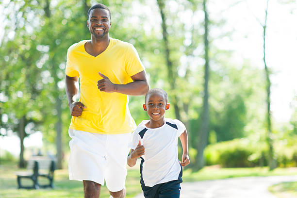 A man and a young boy jog down the street.  The young man has on a yellow top with white shorts.  The young boy has on a white and black top with black shorts.