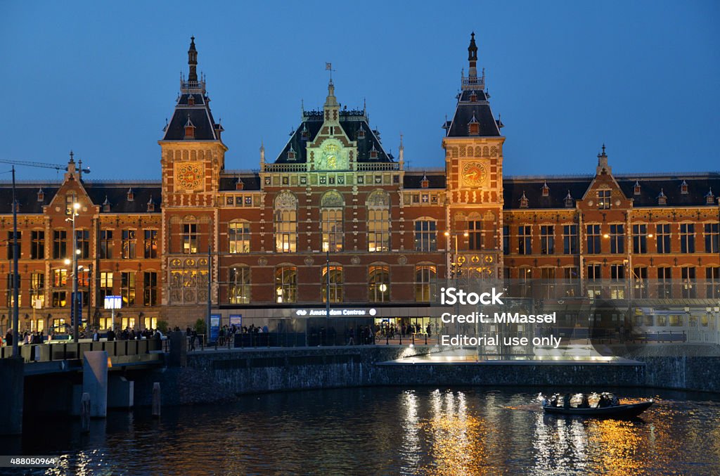 Amsterdam Central Station at night Amsterdam, The Netherlands - September 11, 2015: The main central station external building at night with many people walking around and standing about in front. A tram is also visible. The canal is also shown in foreground and a boat with people inside. Nautical Vessel Stock Photo