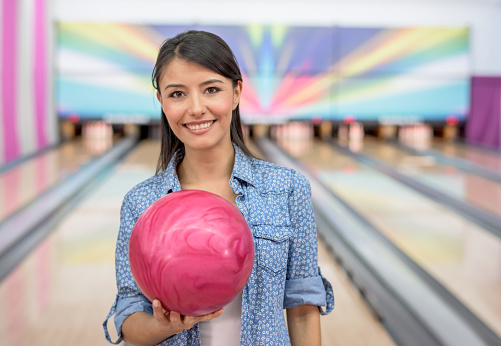 Happy young woman bowling and holding a ball while looking at the camera smiling
