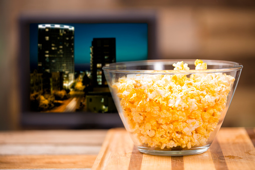 Watching a show or movie on television at home.  Popcorn in clear bowl on coffee table. TV screen with cityscape scene in background.