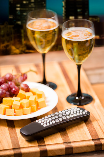 White wine, cheese, red grapes on home coffee table with TV remote control in foreground.  No people. Close up, overhead view. Watching a movie on TV at home with wine and snacks.  City skyline in background. Night.