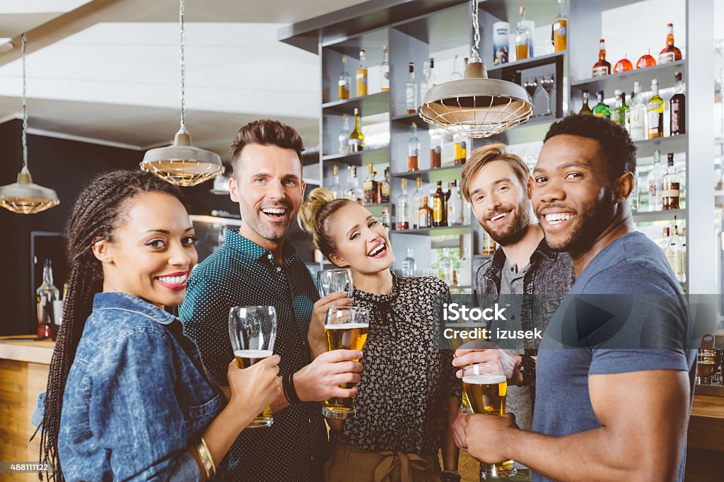 Multi ethnic group of friends drinking beer in a pub Multi ethnic group of happy friends having party in a pub, holding beer glasses and laughing at camera. Bar - Drink Establishment Stock Photo
