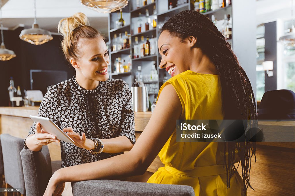 Two young women using a digital tablet Young blonde woman sitting by the counter in the pub and using a digital tablet with her afro american female friend.  2015 Stock Photo