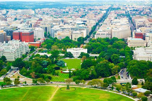 This is an aerial photo showing the National Mall in Washington, D.C. It is a large, open park in downtown Washington, D.C., lined with museums, monuments, and memorials. The United States Capitol can be seen at the far end, serving as a prominent focal point. The National Mall stretches out in a wide grassy expanse with walking paths. Trees line the sides of the Mall, and various cultural institutions, such as the National Museum of American History and the National Gallery of Art, can be seen on either side. The photo is taken from a high vantage point during the daytime, showcasing a clear view under a bright blue sky.