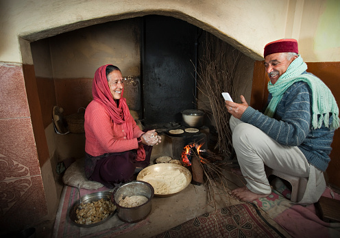 Real people from rural India: Happy senior woman cooking traditional food of Himachal Pradesh called Bereo in her traditional kitchen on bonfire while giving toothy smile to her husband who is holding a mobile phone and reading SMS loudly for her and laughing. Horizontal composition with wide angle lens.